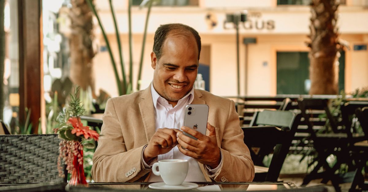 A man in a white shirt smiles while using his smartphone at an outdoor café.