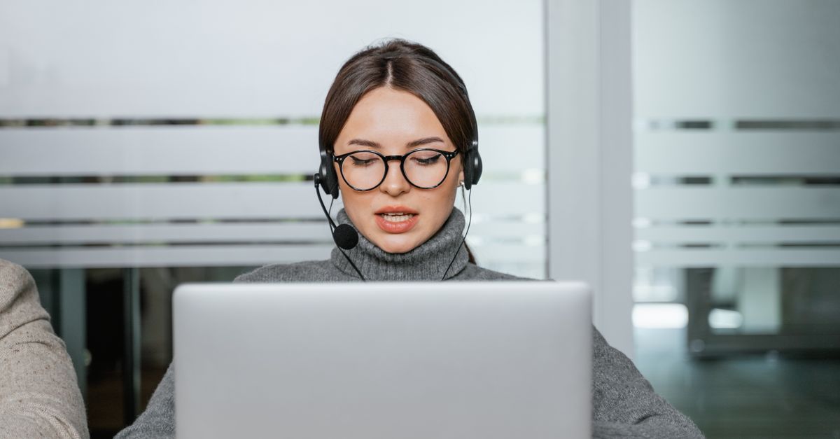 Focused woman with eyeglasses and headset working on laptop in a modern office environment.