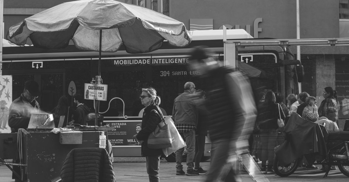 Black and white photo of commuters by a street food stall and bus in the city.